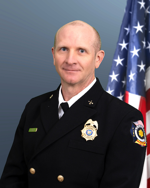 Assistant Fire Chief Dave Wagner in a navy uniform with an American flag in the background against a blue wall