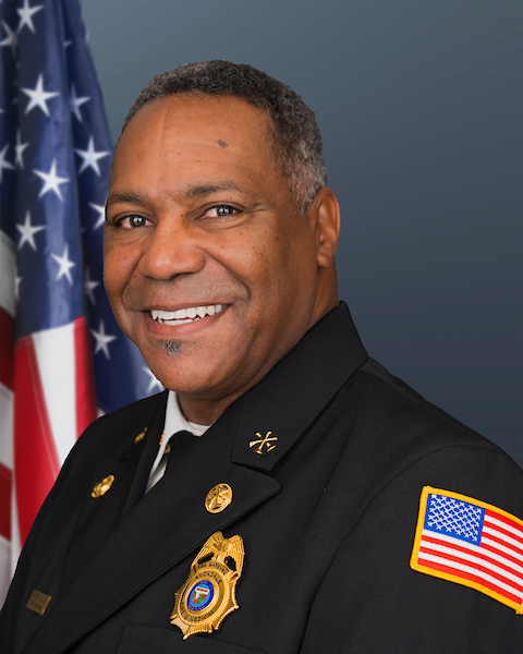 Fire Marshall Scott Miller poses for an official portrait in front of an American flag. He is smiling and dressed in a dark formal dress uniform with department insignia on the sleeve, a gold badge on the chest, and collar pins indicating rank. The background is a plain gradient blue.